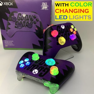 May include: Purple Xbox controller with a Space Jam: A New Legacy design. The controller has a black and white graphic of the words "Goon Squad" and features color-changing LED lights.