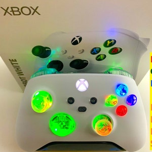 May include: White Xbox controller with illuminated RGB LEDs. The controller has a white body with black buttons and directional pad. The buttons and thumbsticks glow with various colors, including green, blue, and red. The Xbox logo is visible on the controller and the box.