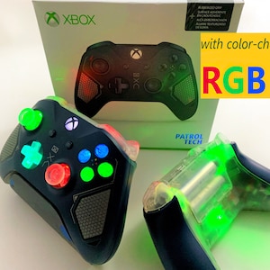 May include: Two black Xbox controllers with RGB LEDs. The controller on the left has green, blue, and red lights. The controller on the right has green lights. The controllers are on a white surface. The box for the controller is in the background. The box is white with the Xbox logo and the text "with color-changing RGB LEDs".
