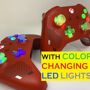 以下が含まれることがあります： 色を変えるLEDライトが付いた赤いXboxコントローラーが2つ。コントローラーの背面には幾何学模様があり、透明なボタンが付いています。