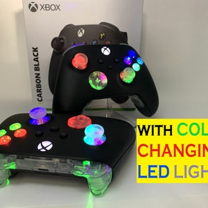 Puede incluir: Dos mandos Xbox negros con detalles en plástico transparente y luces LED de colores. El mando de la izquierda tiene luces verdes, rojas y blancas. El mando de la derecha tiene luces azules, verdes y blancas. El texto "WITH COLOR CHANGING LED LIGHTS" es visible en la imagen.