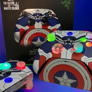 May include: Xbox controller with a Captain America shield design. The controller has a blue, white, and red color scheme, with illuminated buttons. The box in the background has the text "THE FALCON WINTER SOLDIER".