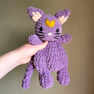 May include: A crocheted purple cat plush toy with a yellow crescent moon on its forehead. The cat has black eyes and a white nose.