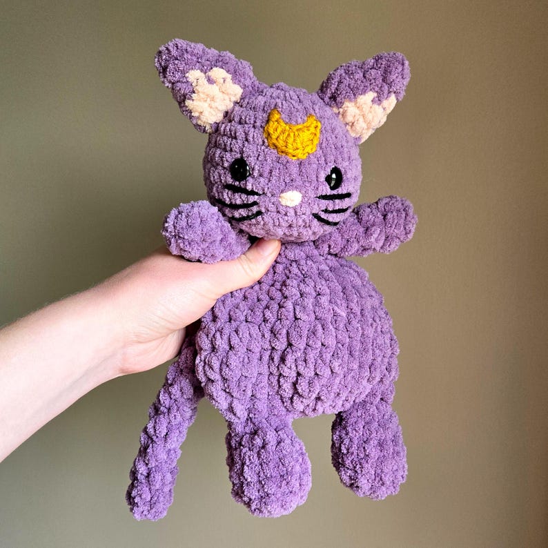 May include: A crocheted purple cat plush toy with a yellow crescent moon on its forehead. The cat has black eyes and a white nose.