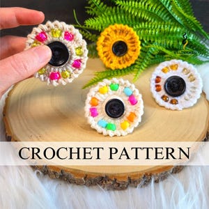 May include: Four circular crochet flower-shaped ornaments featuring a central black button and colourful beads embedded in the yarn. The crochet pieces are white, yellow, and cream-coloured, with the text CROCHET PATTERN displayed across the image.