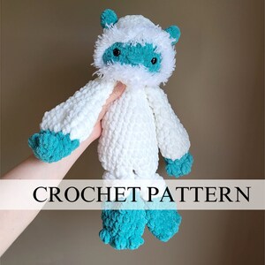 May include: White crocheted yeti plush toy with teal blue accents. The toy has a fluffy white head with a teal blue face and black eyes. The toy is being held by a hand.