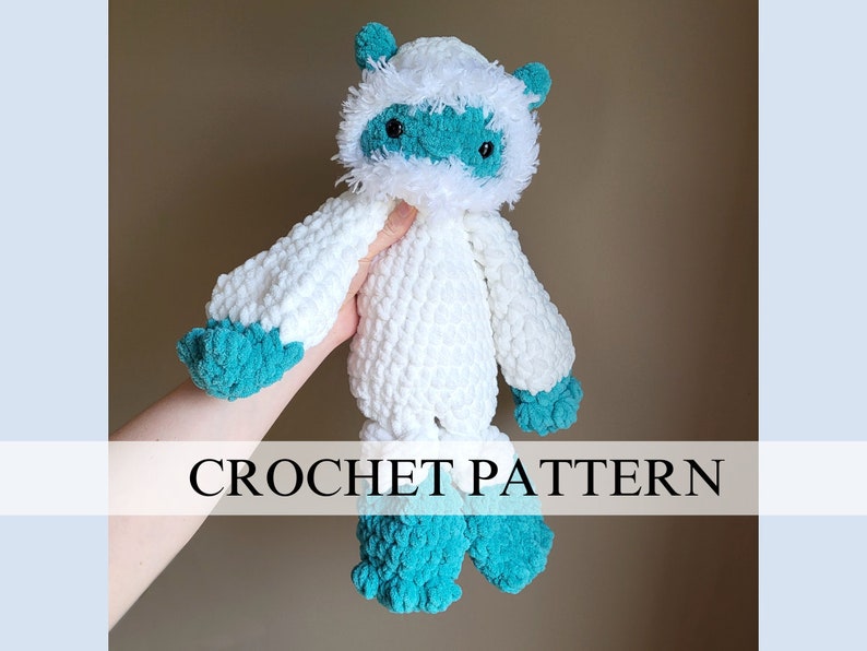May include: White crocheted yeti plush toy with teal blue accents. The toy has a fluffy white head with a teal blue face and black eyes. The toy is being held by a hand.