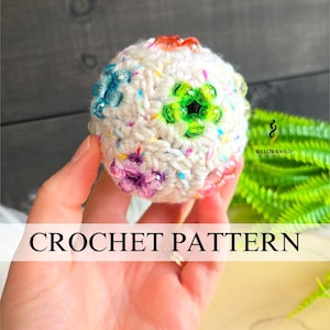 May include: A spherical, off-white crochet ball featuring a speckled yarn texture and embedded with colorful, translucent plastic beads arranged in floral patterns. The text CROCHET PATTERN is displayed prominently across the lower portion of the image.