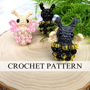 May include: Three handmade crochet bee figures in beige, black, and gray, adorned with beaded wings and bodies. The bees are arranged on a wooden surface with greenery in the background. The image includes the text "CROCHET PATTERN".