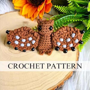 May include: A crocheted bat pattern in shades of brown, with white and black accents. The bat has large, round eyes and is displayed on a wooden surface with a sunflower and greenery in the background. The text "CROCHET PATTERN" is displayed below the bat.