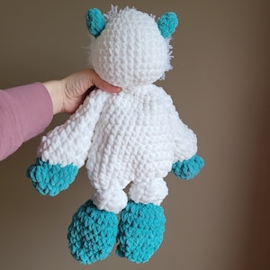 May include: White crocheted plush toy with teal blue accents. The toy has a fluffy white body with teal blue ears, arms, and feet.
