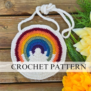 Könnte beinhalten: Eine gehäkelte Regenbogen-Tasche mit einem weißen Trageriemen. Die Tasche ist rund und hat ein Regenbogen-Design mit einer weißen Wolke in der Mitte. Die Tasche liegt auf einer Holzoberfläche mit grünen Pflanzen und gelben Blumen.