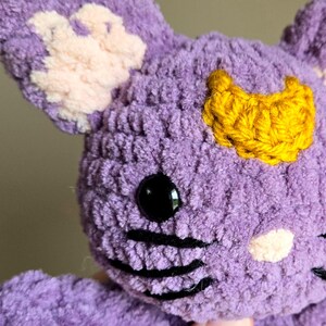 May include: Purple crocheted plush toy with a yellow crescent moon on its head, black embroidered whiskers, and a pink nose.