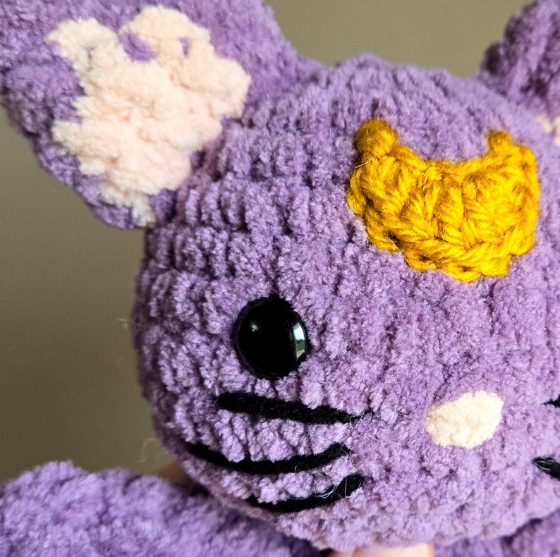 May include: Purple crocheted plush toy with a yellow crescent moon on its head, black embroidered whiskers, and a pink nose.