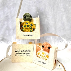 K&ouml;nnte beinhalten: Zwei geh&auml;kelte Schildkr&ouml;ten-Fidget-Spielzeuge in kleinen Geschenkboxen mit einem wei&szlig;en Schleifenbandgriff. Die Boxen sind mit einem maritimen Muster und dem Text "Turtle Fidget" verziert.