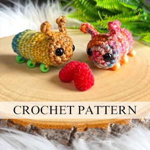 Puede incluir: Dos juguetes de oruga de ganchillo con ojos negros grandes y antenas pequeñas. Uno es amarillo, azul y verde, el otro rosa, naranja y morado. Una frambuesa roja se encuentra entre ellos. El texto "CROCHET PATTERN" está en la parte inferior.