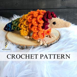 Op de afbeelding: Een kleurrijke gehaakte pangolin met een regenboogstaart en oranje, gele en rode schubben. De pangolin wordt getoond op een houten schijf bovenop een bed van witte imitatiebont. De tekst "CROCHET PATTERN" staat eronder.