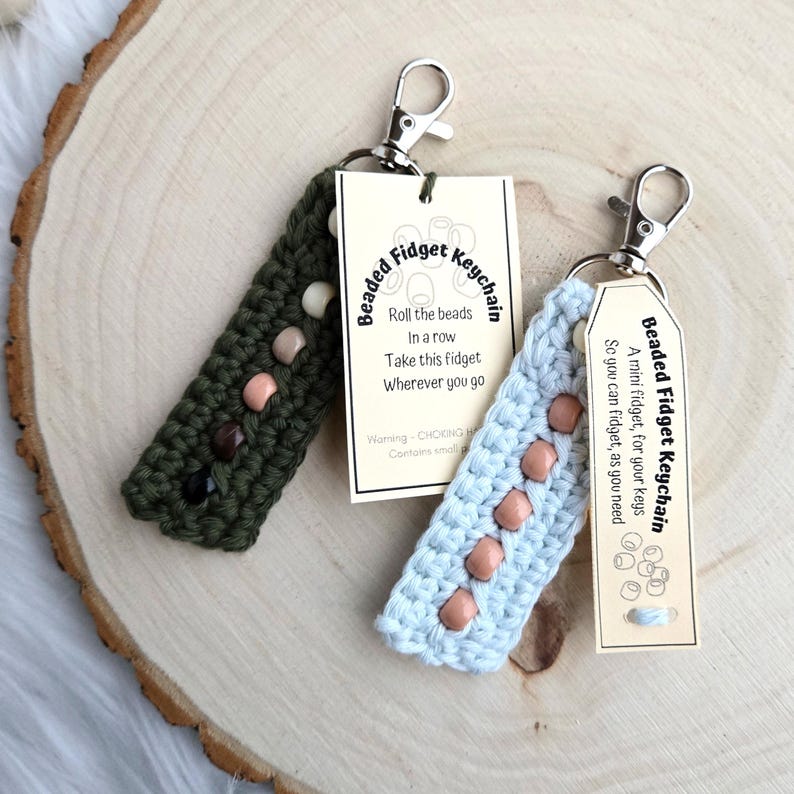 May include: Two crocheted beaded fidget keychains. One is green with brown beads and the other is light blue with peach beads. Both have a metal keychain attached. The tags say "Beaded Fidget Keychain" and include instructions for using the keychain.