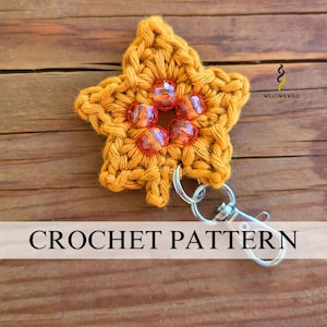Peut inclure: Une étoile au crochet jaune moutarde avec cinq perles orange translucides disposées en cercle au centre. L'étoile est fixée à un mousqueton en métal argenté. Le texte WILLOW & WILD est visible dans le coin supérieur droit.