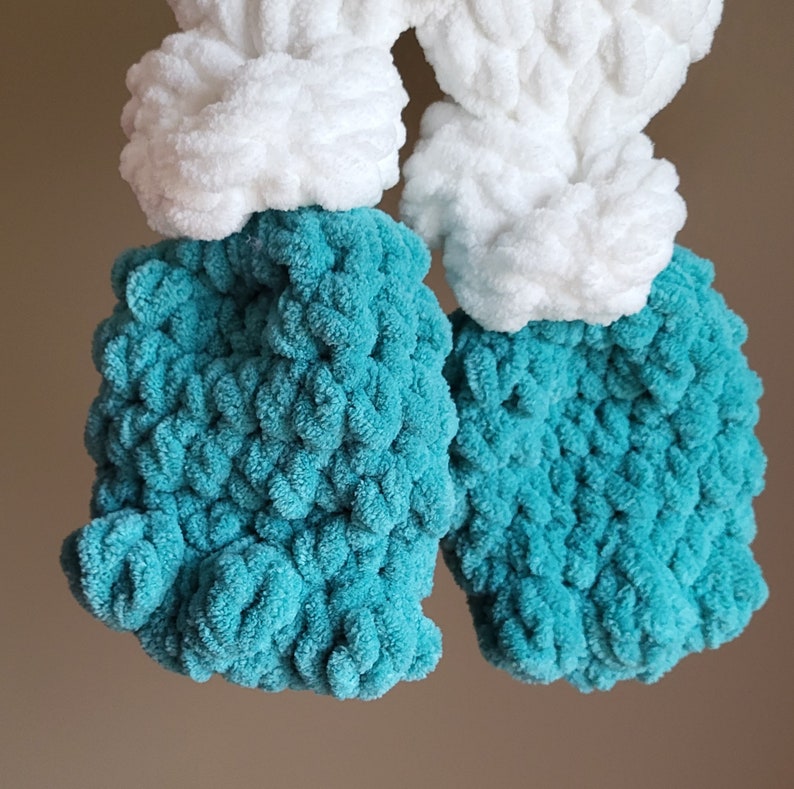 May include: Two crocheted yarn pom-poms, one teal and one white. The teal pom-pom is on the left and the white pom-pom is on the right.
