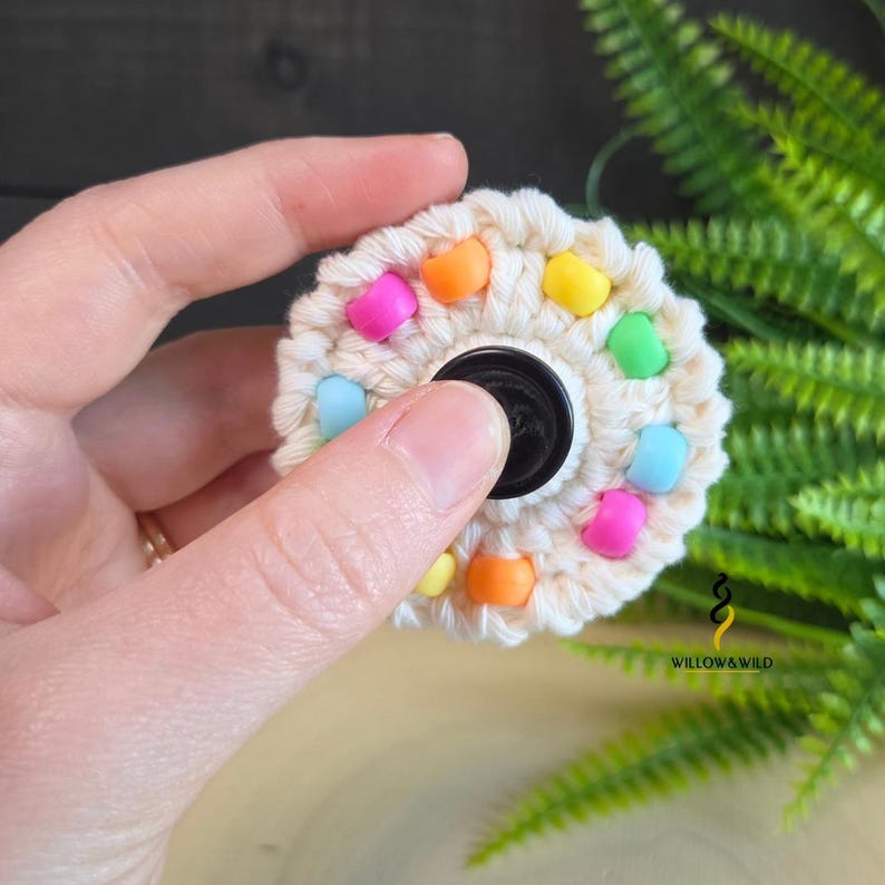 May include: A circular fidget spinner toy featuring a cream-coloured crocheted yarn exterior adorned with small, vibrant plastic beads in shades of orange, yellow, pink, and light blue, centred around a smooth black circular bearing component.