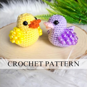May include: Two crocheted ducklings, one yellow with orange beak, the other purple with pink beak, sit on a wooden surface. The yellow duckling has yellow bead accents. The image includes the text "CROCHET PATTERN".