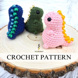 May include: Three crocheted dinosaur toys on a wooden slice. The dinosaurs are blue, green, and pink. The blue dinosaur has a blue and green beaded tail. The green dinosaur has a gold beaded head. The pink dinosaur has a yellow beaded tail. The text "CROCHET PATTERN" is printed on the wooden slice.