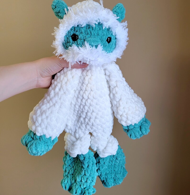 May include: A crocheted plush toy in the shape of a yeti. The toy is white with a teal blue face and arms and legs. The toy has a fluffy white beard and a teal blue nose.