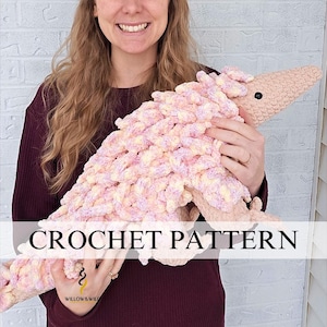 Può includere: Un grande dinosauro all'uncinetto fatto a mano, nei toni del rosa e del crema, è tenuto da una persona. Il dinosauro ha un aspetto strutturato e soffice. Il testo "CROCHET PATTERN" è visualizzato sull'immagine. La testa del dinosauro è di colore beige chiaro.