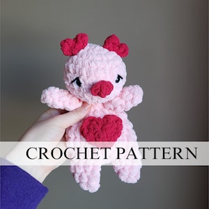 May include: A pink crocheted amigurumi toy with a red heart on its chest. The toy has a heart-shaped nose and ears. The toy is being held by a hand.