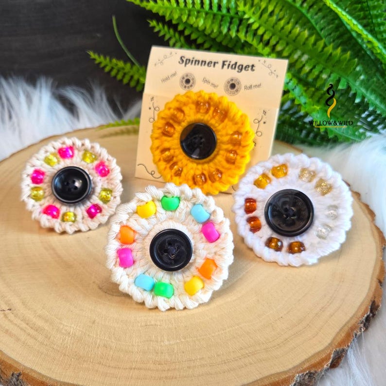 May include: Three circular crochet spinner fidget toys with black buttons in the centre and colourful beads around the edges. One is yellow, one is cream with neon beads, and one is cream with amber beads. A display card reads Spinner Fidget.