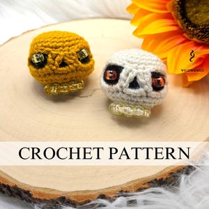 May include: Two crocheted skull ornaments, one yellow and one white, with black eye sockets and orange bead eyes. The skulls sit on a wooden slice, with a sunflower in the background. The text "CROCHET PATTERN" is displayed.