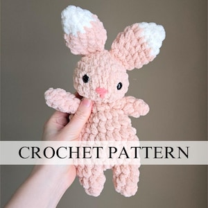 May include: A handmade, pink crochet bunny with white-tipped ears, black eyes, and a pink nose. The bunny is held in a hand. The text "CROCHET PATTERN" is displayed across the image.