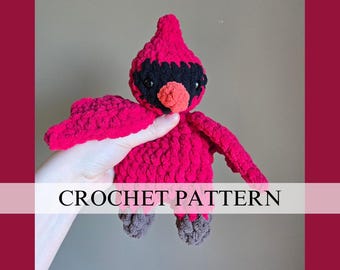 May include: A handmade, red crochet cardinal bird with black accents and an orange beak. The bird has outstretched wings and gray feet. The text "CROCHET PATTERN" is displayed across the image.