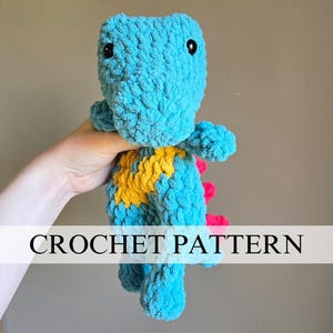 May include: A handmade, turquoise crochet dinosaur with black eyes, yellow and pink accents. The text "CROCHET PATTERN" is displayed across the image. This plush toy is a unique, handcrafted item.