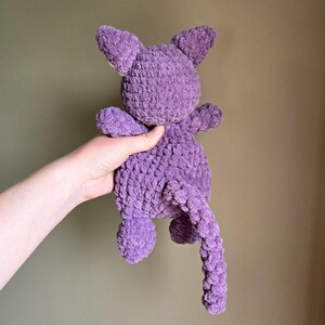 May include: A purple crocheted stuffed animal in the shape of a cat. The cat has a long tail and is being held by a hand.