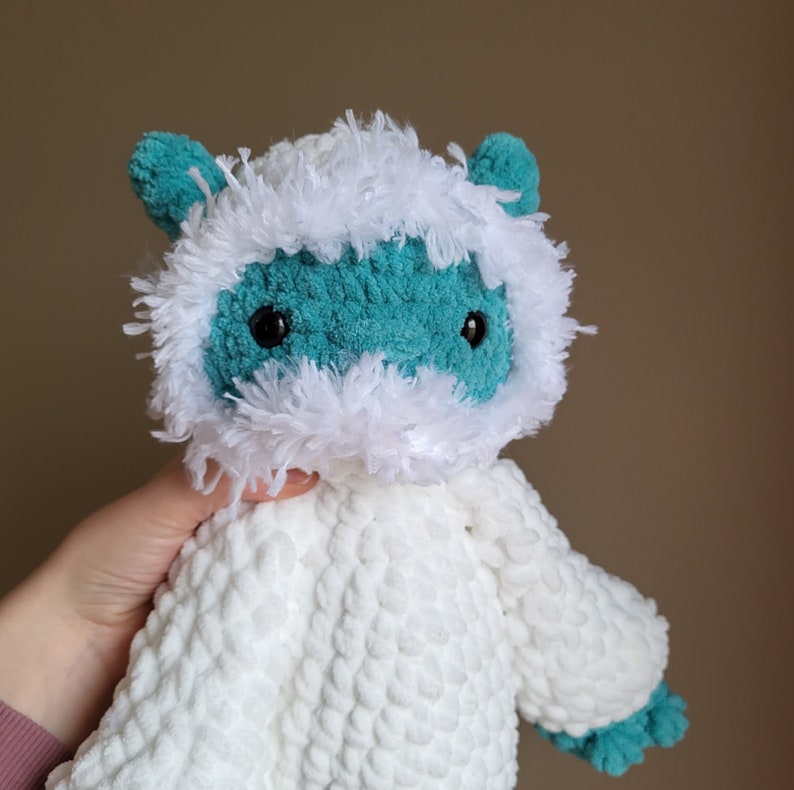 May include: A crocheted plush toy with a white fur coat and a teal blue head. The toy has black eyes and a white fur trim around the face.