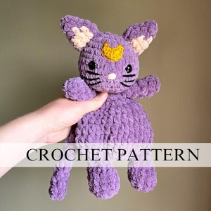 May include: A crocheted purple cat toy with a yellow crescent moon on its head. The cat has black eyes and a black nose. The toy is being held by a hand.