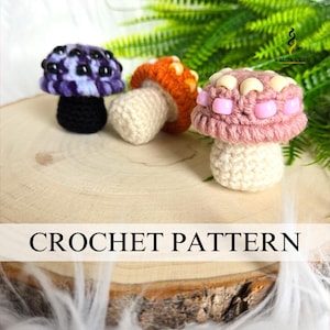 May include: Three handmade crochet mushroom figures in purple, orange, and pink, with white stems, sit on a wooden slice. The mushrooms have textured caps and are displayed against a backdrop of green foliage. The text "CROCHET PATTERN" is visible.