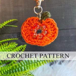 May include: A crocheted orange pumpkin keychain featuring a small green stem and a center ring adorned with five clear, faceted beads. The pumpkin is attached to a silver-toned metal lobster clasp and split ring. The text CROCHET PATTERN is visible.