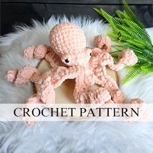 Può includere: Un giocattolo a forma di polpo all'uncinetto rosa chiaro fatto a mano, con occhi neri. Il polpo è appoggiato su una fetta di legno e finta pelliccia bianca. Il testo "CROCHET PATTERN" è visibile.