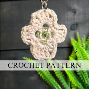 May include: A crocheted flower-shaped keychain in a light beige color, adorned with clear beads at the center. The text "CROCHET PATTERN" is displayed on a banner across the image. The background features dark wood and green foliage.
