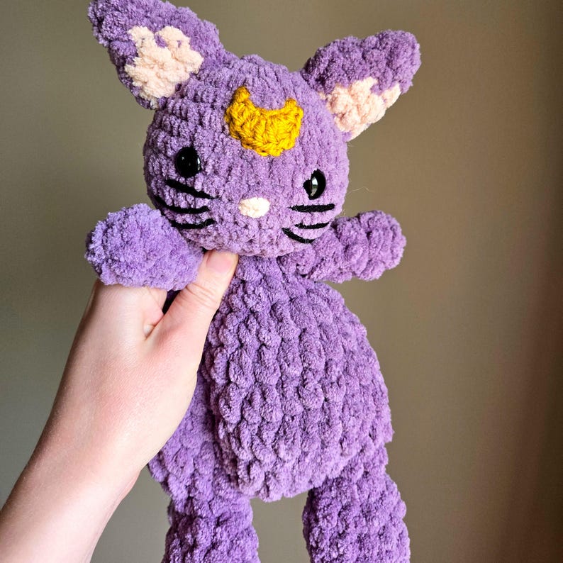 May include: A purple crocheted stuffed animal shaped like a cat with a yellow crescent moon on its forehead. The cat has black eyes and a black stitched nose and whiskers.