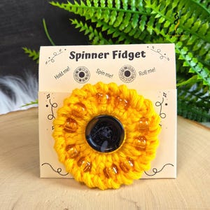 May include: A yellow crocheted spinner fidget toy with amber-coloured beads attached to a beige display card. The card features the text Spinner Fidget, Hold me!, Spin me!, and Roll me! with decorative line art and the Willow & Wild logo.