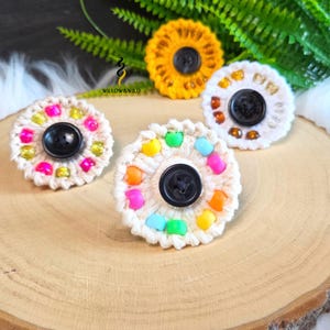 May include: Three circular crochet ornaments featuring a central black button surrounded by colourful beads. One is yellow, one is white with amber beads, and one is white with neon beads. The text WILLOW&WILD is printed in black above the yellow ornament.