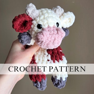 May include: Crochet pattern for a plush cow toy. The cow is white with red, pink, and gray accents. The toy is being held by a hand.