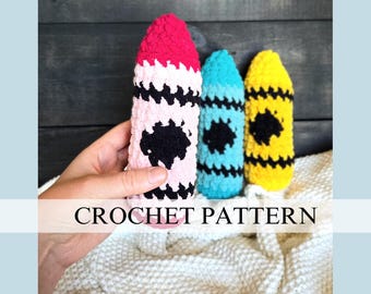 May include: Three crocheted crayon toys in pink, blue, and yellow with black detailing.  The crayons are sitting on a white crocheted blanket. CROCHET PATTERN is written in black text on a white banner.
