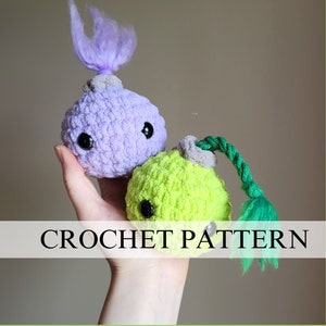 May include: Two crocheted plush toys, one purple with a purple yarn tail and one green with a green yarn tail. The toys have black button eyes and are held in a person's hand. CROCHET PATTERN is written in white text on a white banner.
