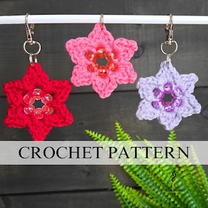 May include: Three handmade crochet flower keychains in red, pink, and lavender, each with a beaded center. The keychains are suspended from a white bar against a dark wood background. The image also includes the text "CROCHET PATTERN".