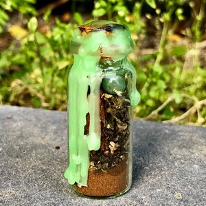 May include: A small glass bottle filled with layers of herbs, spices, and a small green stone. The bottle is sealed with a cork and green wax drippings. The background is blurred green foliage.